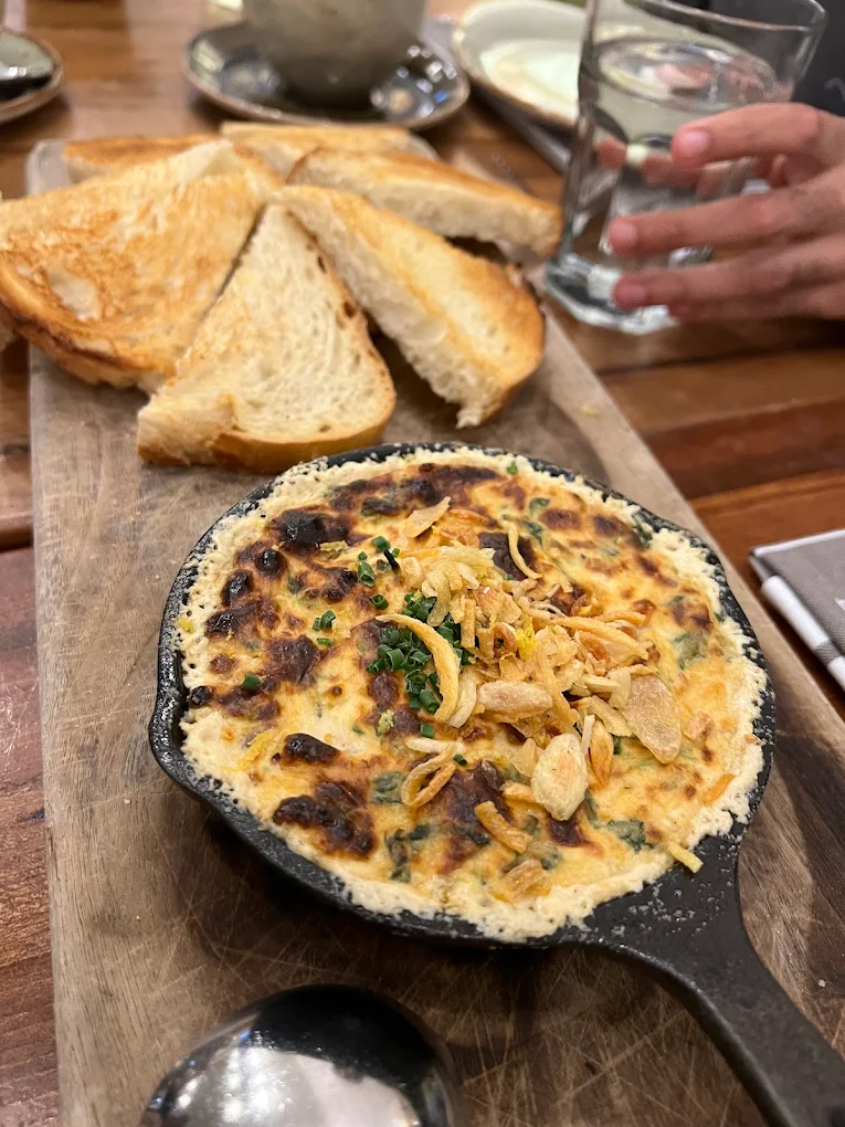 A small cast iron skillet filled with a creamy, baked dip topped with herbs and crispy onions sits on a wooden board next to slices of toasted bread; a hand reaches for a glass of water in the background.