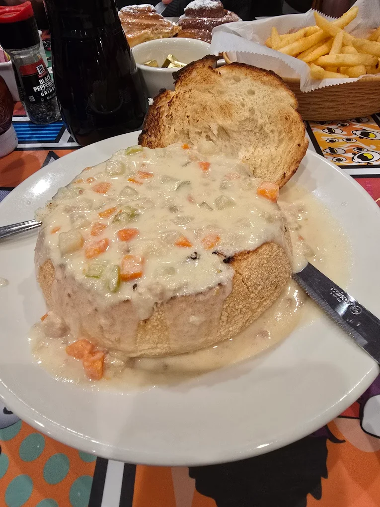 A bread bowl filled with creamy soup containing diced carrots, celery, and potatoes, served with a toasted bread slice on a white plate; fries and other food items are visible in the background.