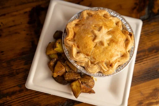 A golden-brown pot pie with a decorative star crust sits on a white square plate, accompanied by a side of roasted potatoes, on a wooden table.