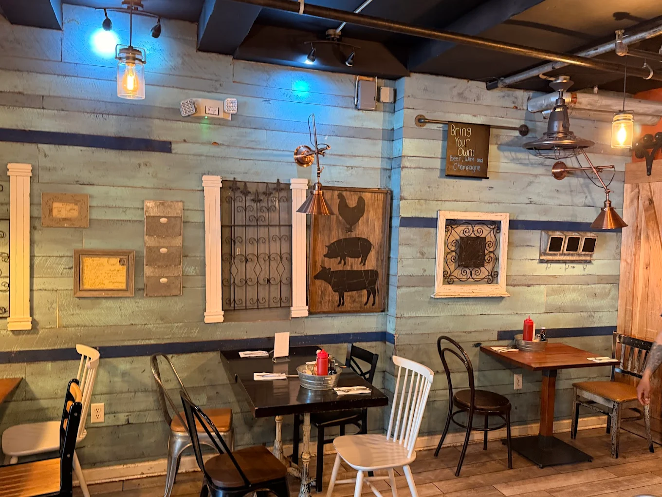 A cozy restaurant with rustic blue wooden walls, eclectic wall art including farm animal silhouettes and metal grates, and mismatched chairs around wooden tables set with condiments. Warm lighting adds to the inviting atmosphere.