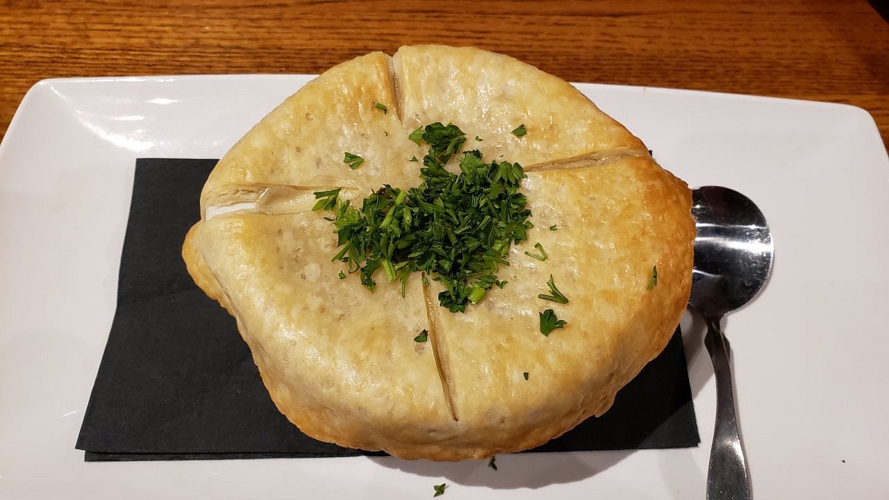A bowl of pot pie topped with a golden, flaky pastry crust, garnished with chopped parsley, sits on a white plate with a black napkin and a spoon beside it.