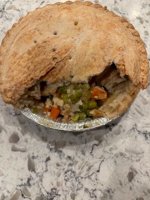 A partially eaten pot pie with a golden-brown crust sits on a marble countertop, revealing a filling of chicken, peas, and carrots.