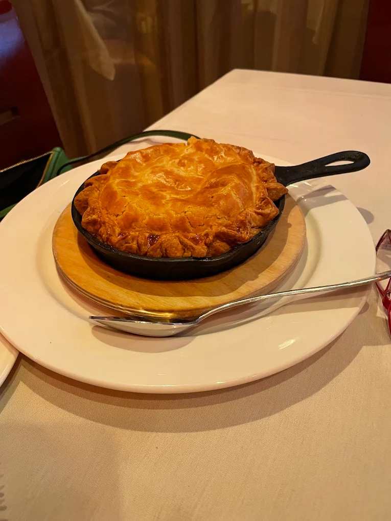 A golden-brown pie with a flaky crust sits in a small cast iron skillet on a wooden trivet, placed on a white plate with a spoon beside it on a table covered with a white tablecloth.