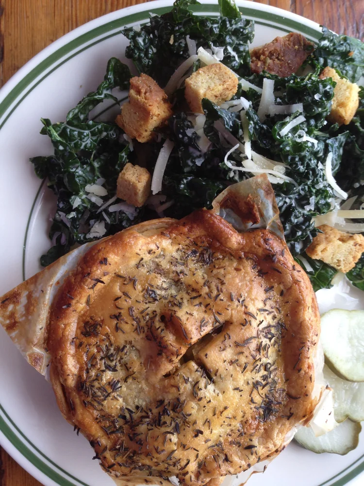 A savory herb-crusted pot pie on a white plate with green trim, served alongside a kale salad with croutons, shredded cheese, and a few cucumber slices.