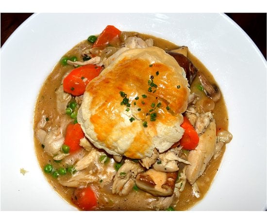 A white bowl filled with chicken pot pie, featuring chunks of chicken, carrots, peas, and gravy, topped with a golden, flaky biscuit and a sprinkle of chopped chives.