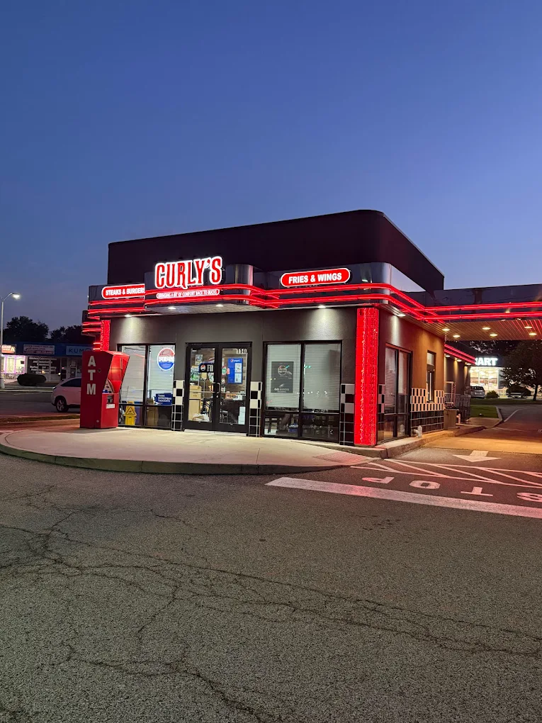 A brightly lit Curly’s restaurant at dusk features red neon lights and signage for fries and wings; an ATM stands near the entrance, and the parking lot is mostly empty under a clear evening sky.