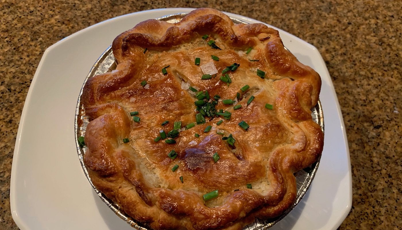 A golden-brown pot pie topped with chopped green herbs sits on a white plate, placed on a speckled brown countertop.