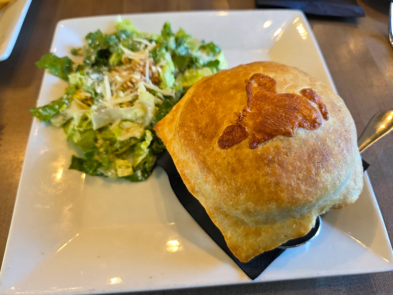 A square white plate with a leafy green Caesar salad topped with shredded cheese on the left and a golden-brown pastry with a baked butterfly shape on top on the right.