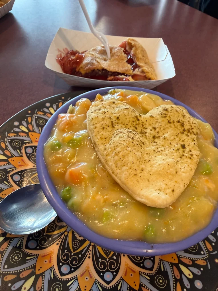 A bowl of pot pie with a heart-shaped biscuit on top, served on a decorative plate beside a spoon. In the background, a serving of berry pie with a fork rests in a paper tray.