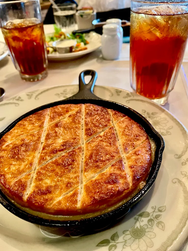 A golden-brown savory pie with a crisscross pattern is served in a small cast iron skillet, alongside glasses of iced tea and plates of salad on a tabletop.