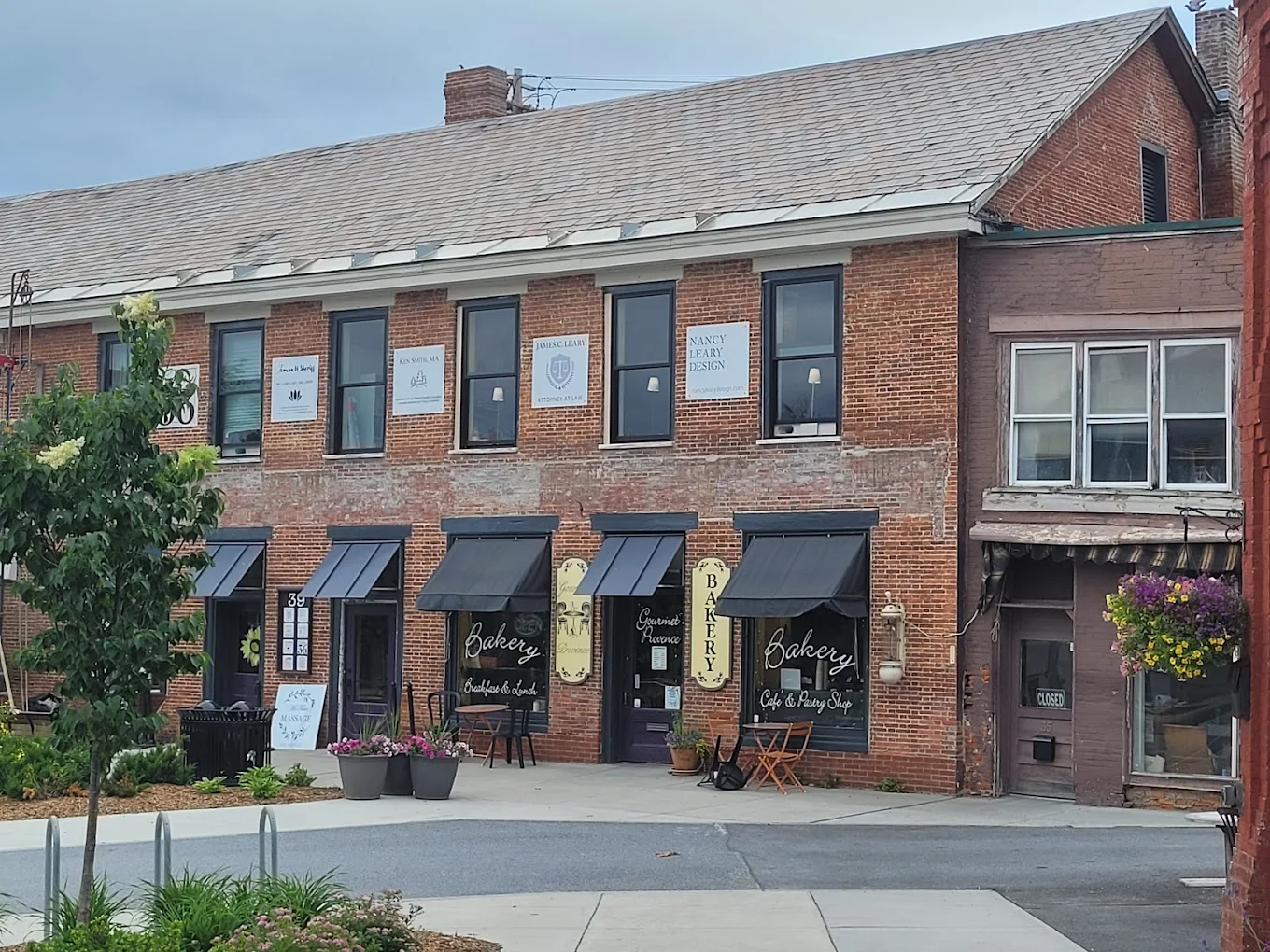 A row of brick buildings with black awnings houses a bakery and other businesses. Outdoor tables and potted plants decorate the sidewalk, and a hanging flower basket is visible on the right. The sky is overcast.