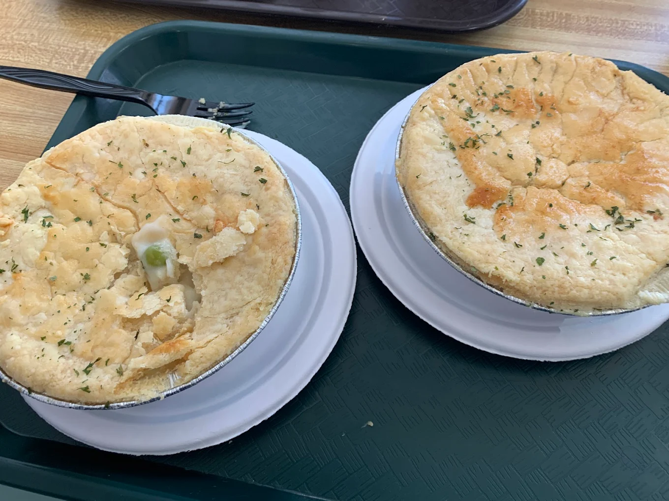 Two individual chicken pot pies with golden, flaky crusts are served in metal tins on white plates atop a green cafeteria tray; one pie has a piece of crust broken, revealing peas and creamy filling inside.
