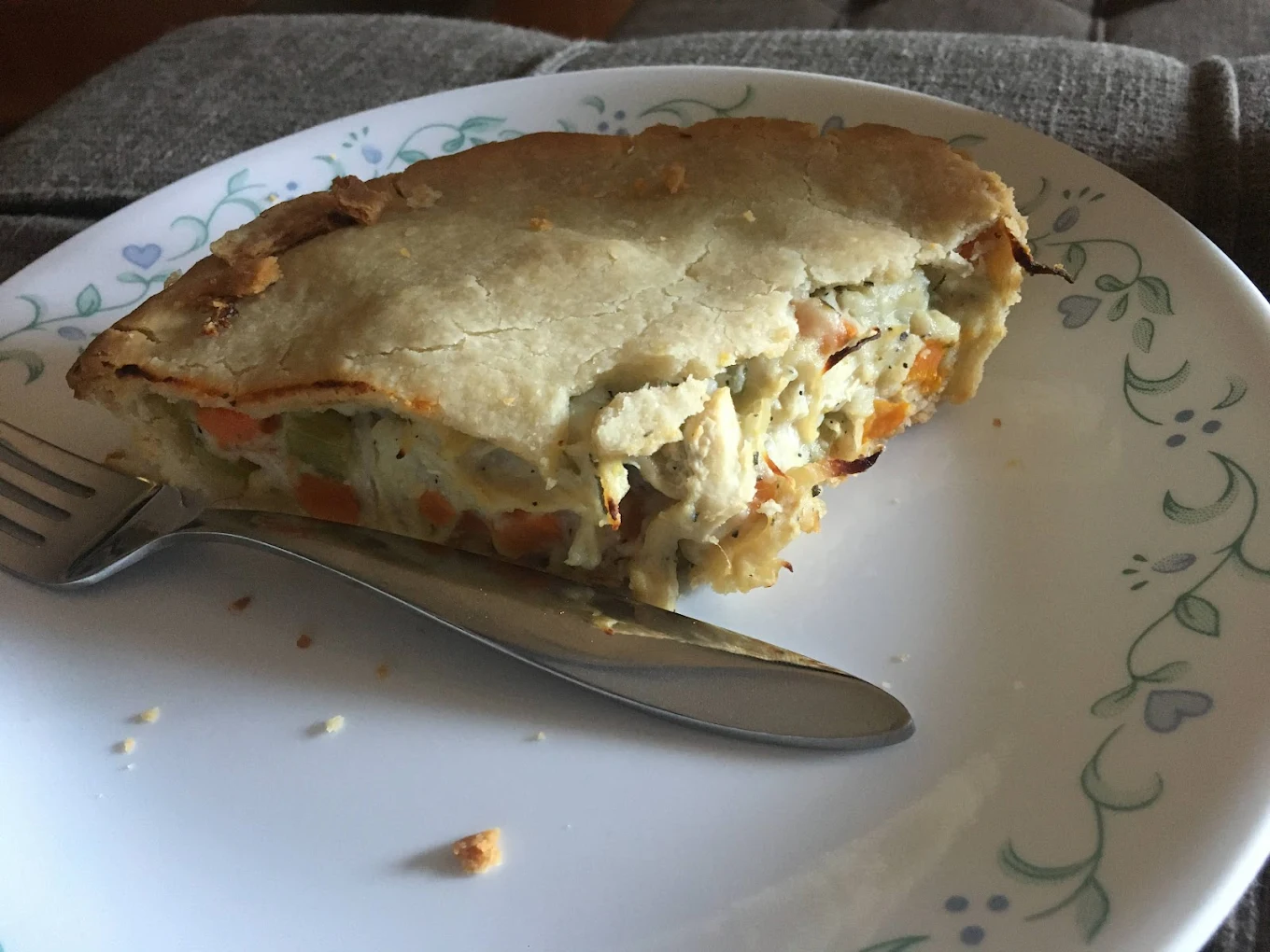 A slice of savory pie with a golden crust and a filling of vegetables and meat sits on a white plate with a floral pattern; a fork and knife rest beside it.