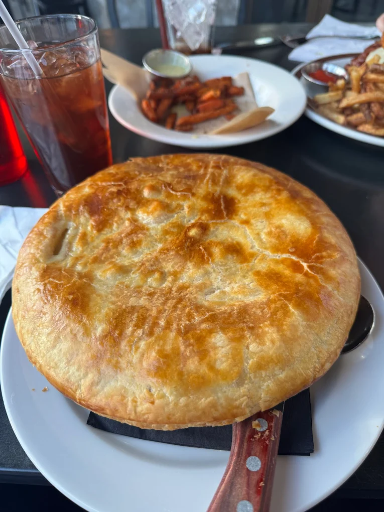 A golden-brown pot pie sits on a white plate with a wooden-handled spoon beside it; in the background are plates of sweet potato fries, regular fries, and a glass of iced tea.