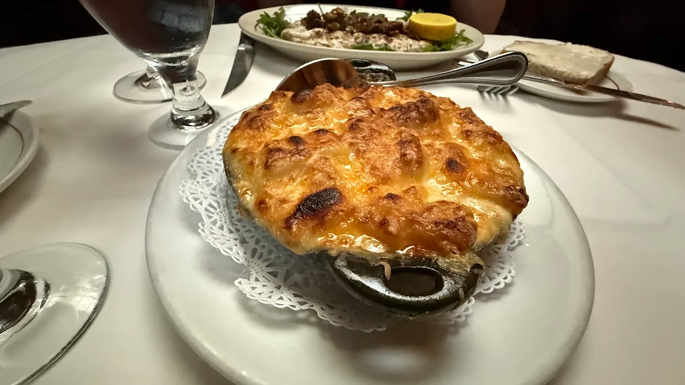 A casserole dish of baked macaroni and cheese with a golden, bubbly crust sits on a white plate with a decorative doily, alongside a glass of water and another dish in the background on a restaurant table.