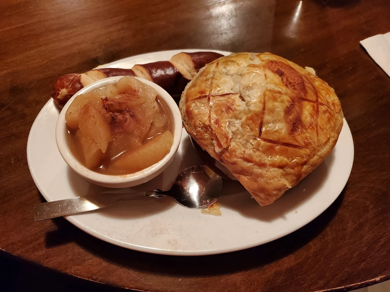 A white plate with a bowl of cooked apples, a spoon, a piece of pretzel bread, and a golden-brown puff pastry pie on a wooden table.