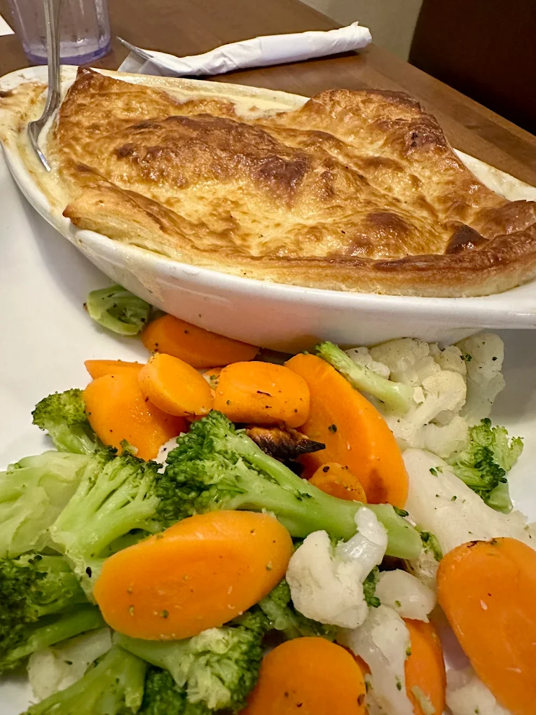 A baked pot pie with a golden-brown crust is served in a white dish, accompanied by steamed broccoli, carrots, and cauliflower on a white plate. A folded napkin and glass are in the background.
