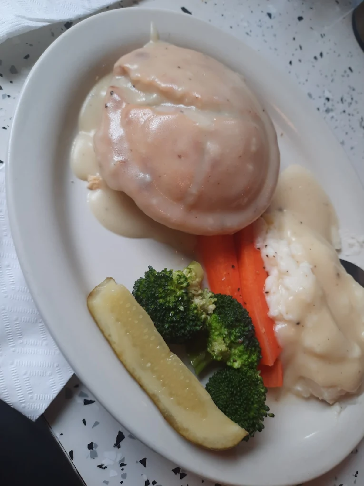 A plate with mashed potatoes and brown gravy, steamed broccoli and carrots, a pickle spear, and a side of white gravy. A napkin is partially visible to the left.