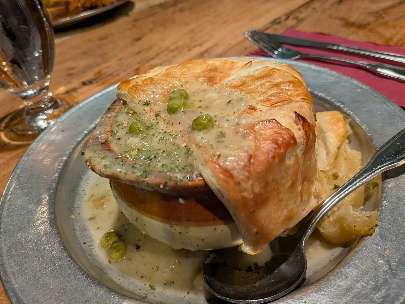 A pot pie topped with golden, flaky pastry sits on a metal plate. Creamy filling with peas spills out, and a spoon rests beside it. A glass, utensils, and a red napkin are on the wooden table.