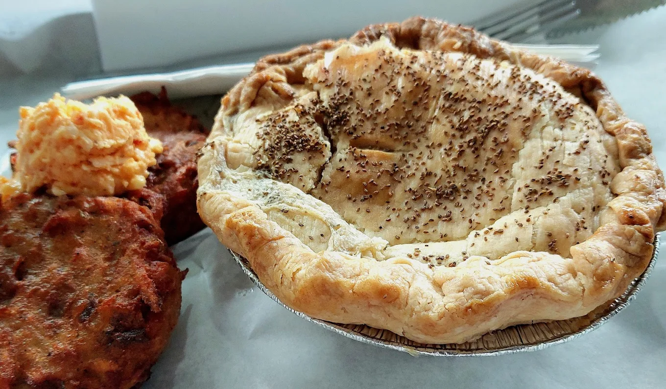 A close-up of a baked savory pie with a flaky, golden-brown crust sprinkled with pepper, served with a side of pimento cheese and a fried patty on a white paper-lined tray.