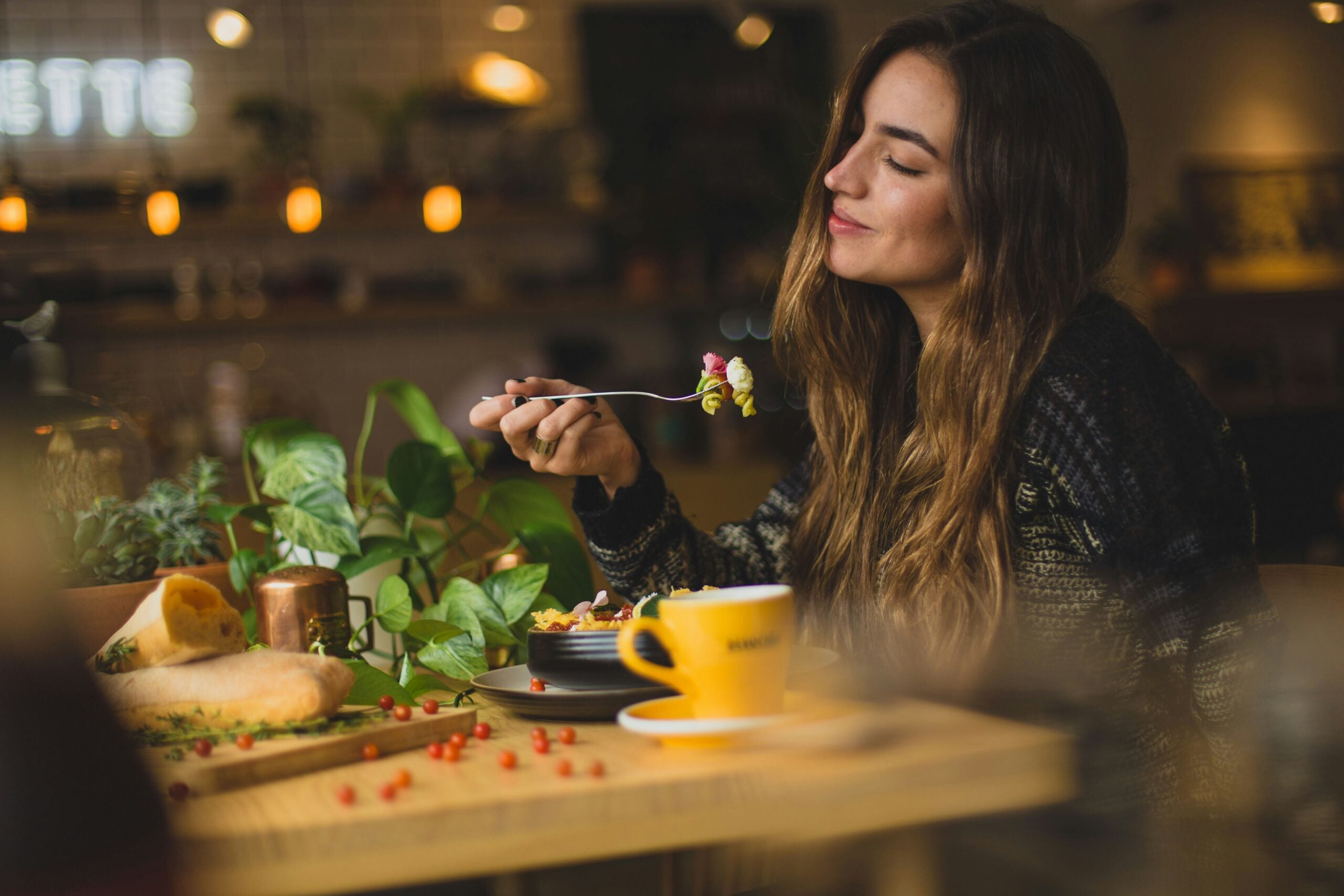 A woman with long brown hair sits at a cozy café table, smiling with her eyes closed as she lifts a forkful of food. A yellow coffee cup, a plate, and green plants are on the wooden table in front of her.