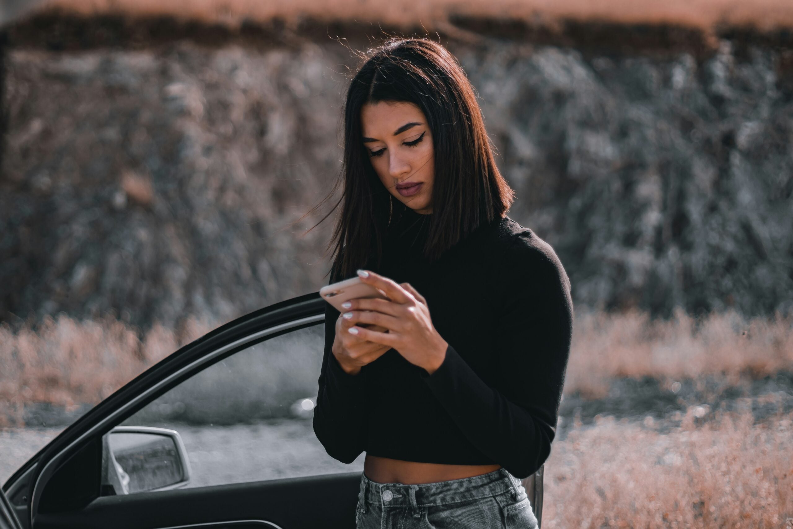 A woman with straight dark hair, wearing a black long-sleeve top and jeans, stands next to an open car door outdoors, looking down at and using her smartphone. The background is blurred and natural.