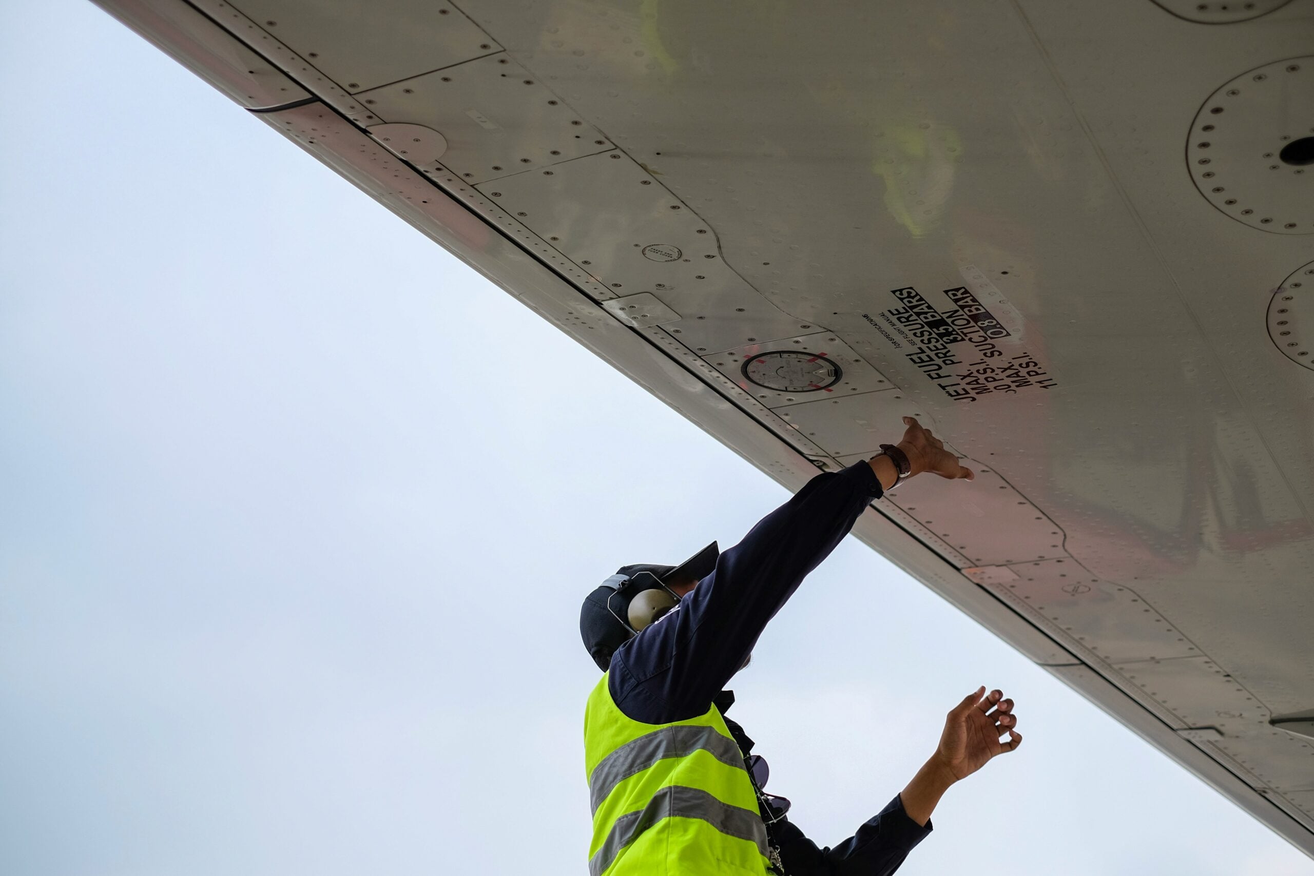 A worker in a yellow safety vest and ear protection inspects or checks under the wing of an aircraft, reaching up toward maintenance markings on the metal surface.