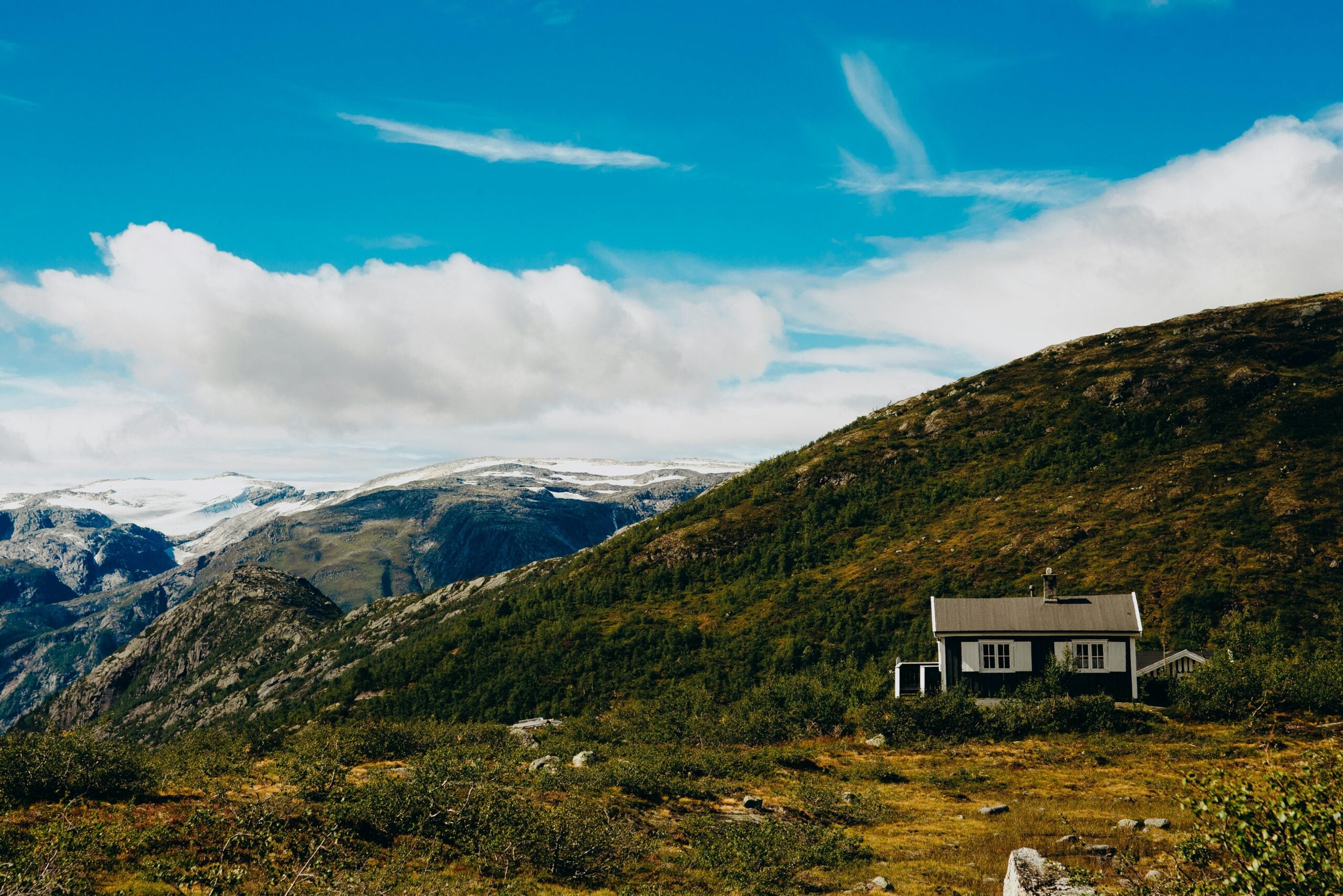 A small white house sits on a grassy hillside with a backdrop of mountains under a blue sky with scattered clouds. The scene is peaceful and surrounded by nature.