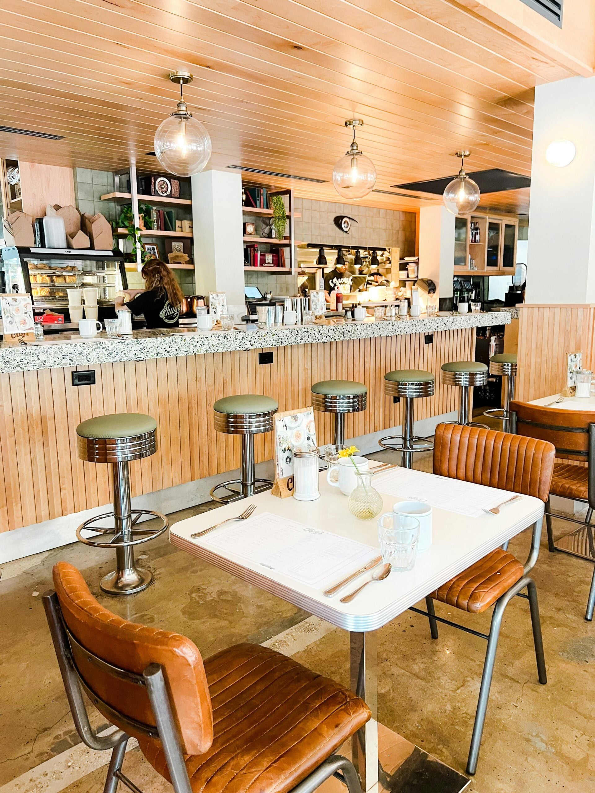 A cozy café with brown leather chairs, a white table set for two, and a counter with metal stools. Shelves, lights, and a barista working behind the counter give the space a warm, inviting atmosphere.