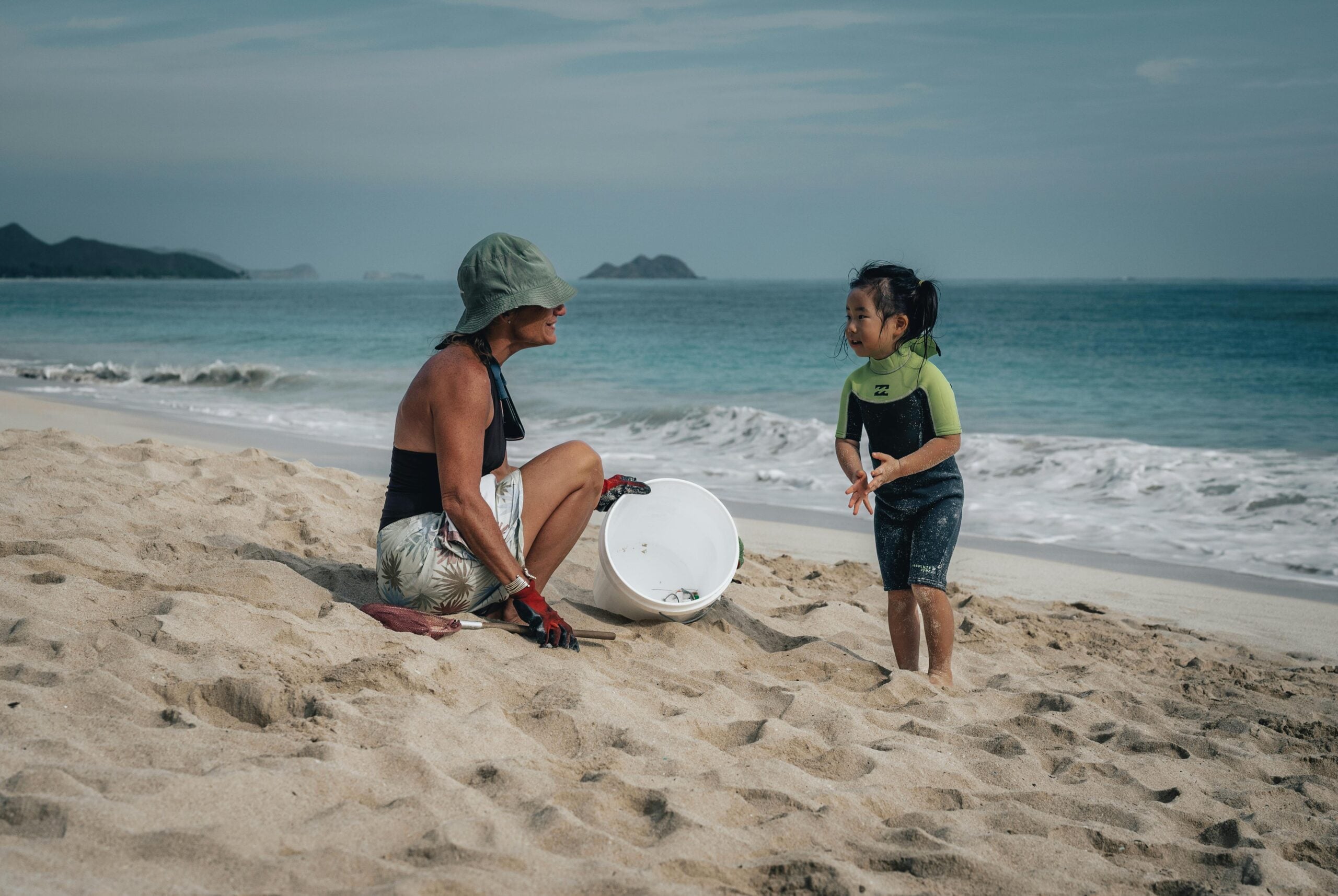An adult and a young child, both in beachwear, smile at each other on a sandy beach near the ocean. The adult is sitting with a white bucket, while the child stands nearby in a wetsuit top.