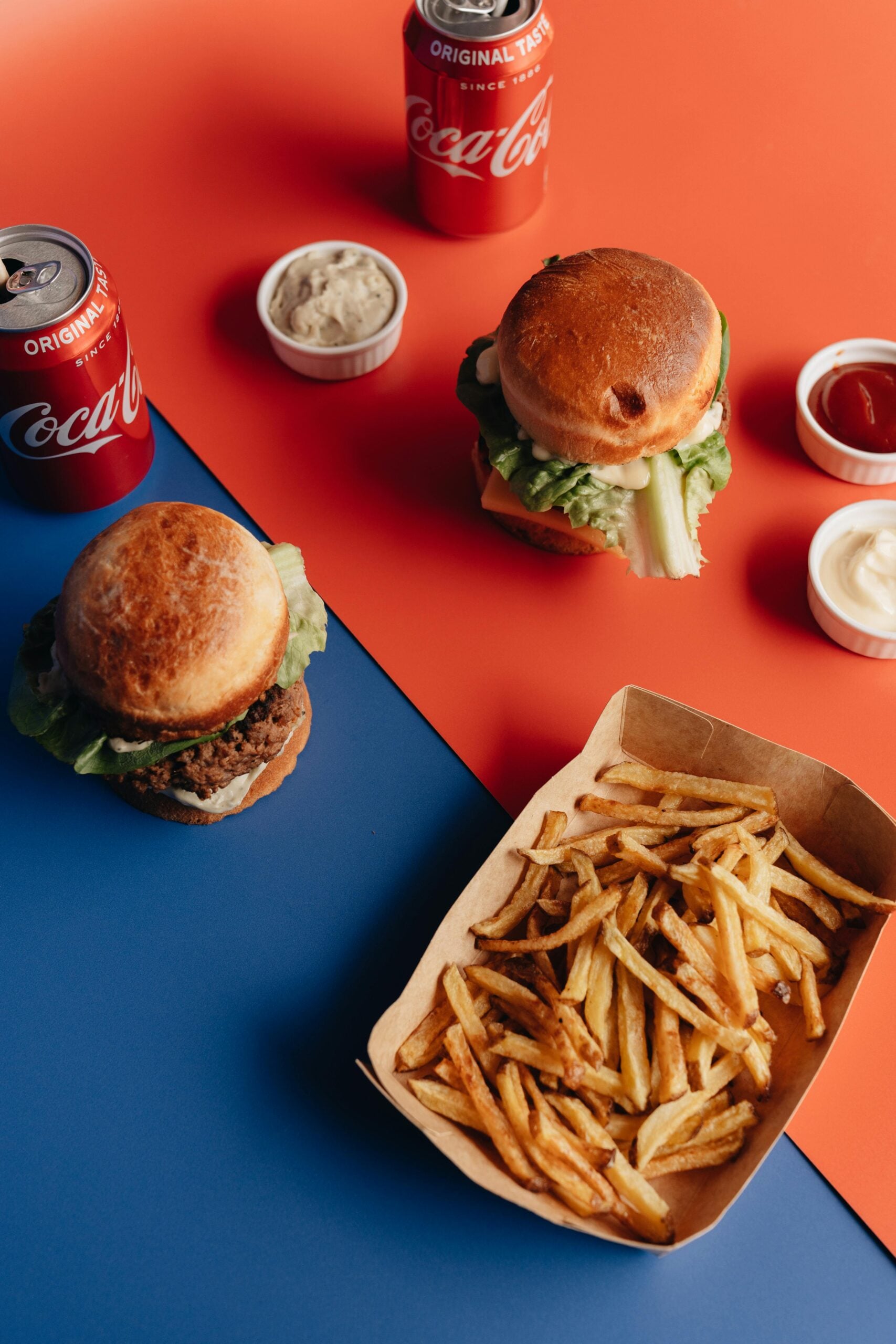 Two burgers with lettuce, a tray of French fries, two cans of Coca-Cola, and small cups of dipping sauces are arranged on a split blue and orange background.