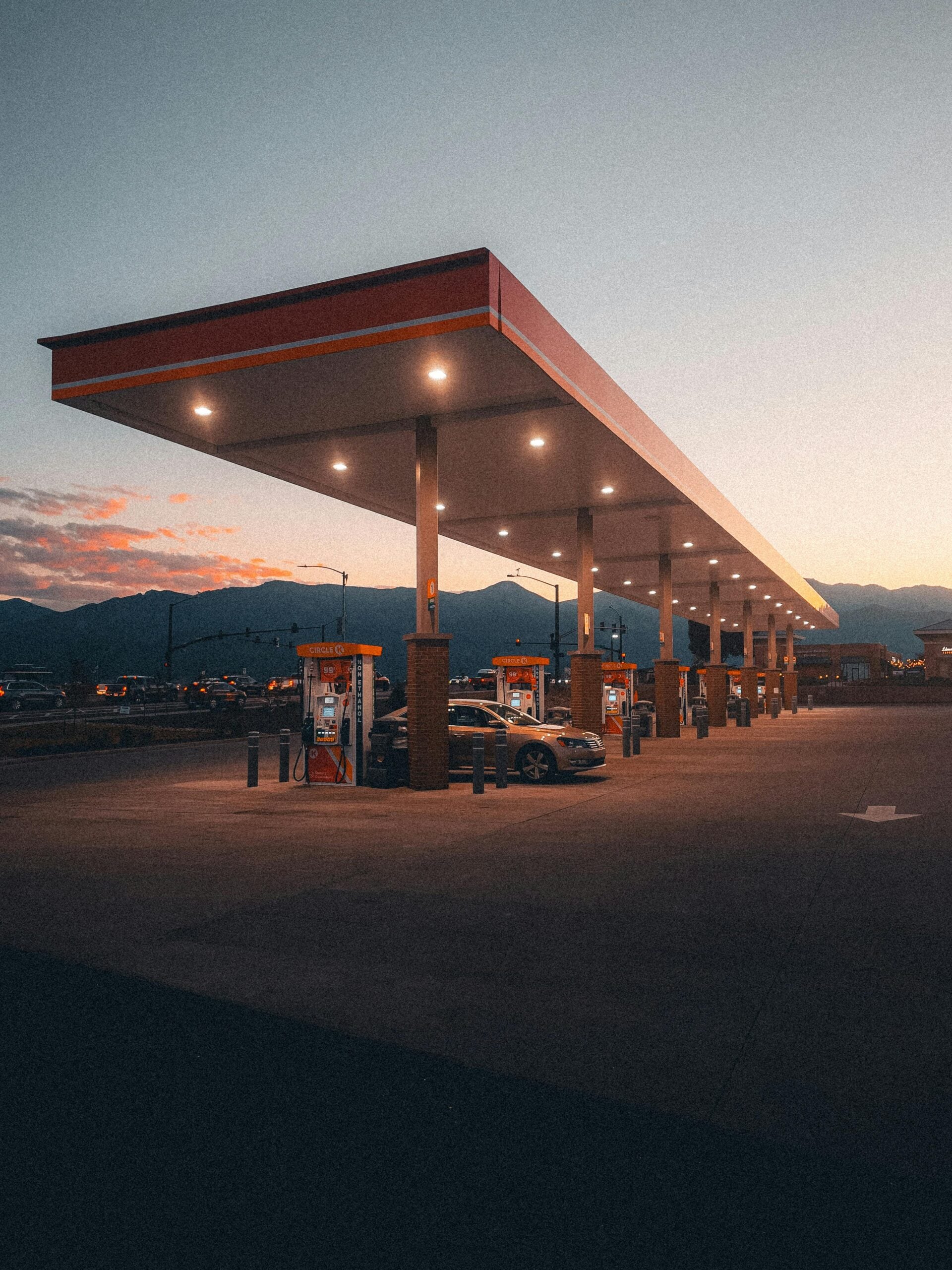 A mostly empty gas station at dusk with a few cars refueling under a long, brightly lit canopy; mountains and a cloudy sky are visible in the background.