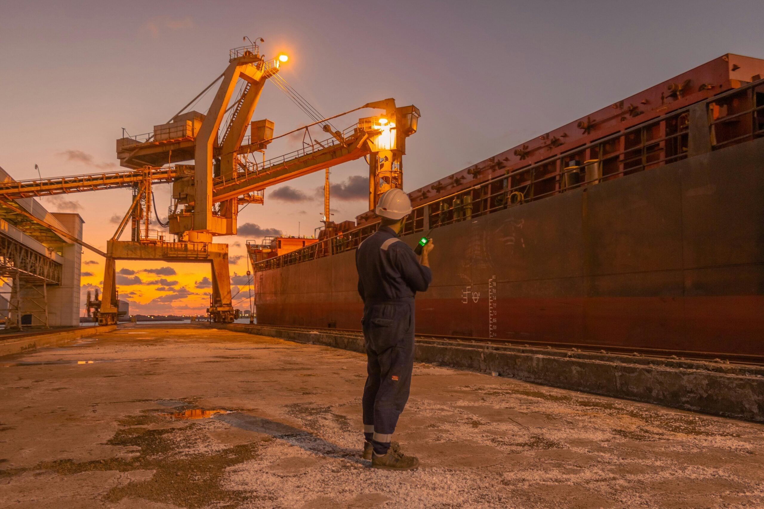 A worker in a helmet and coveralls stands on a dock at sunset, using a walkie-talkie near a large cargo ship and industrial cranes.