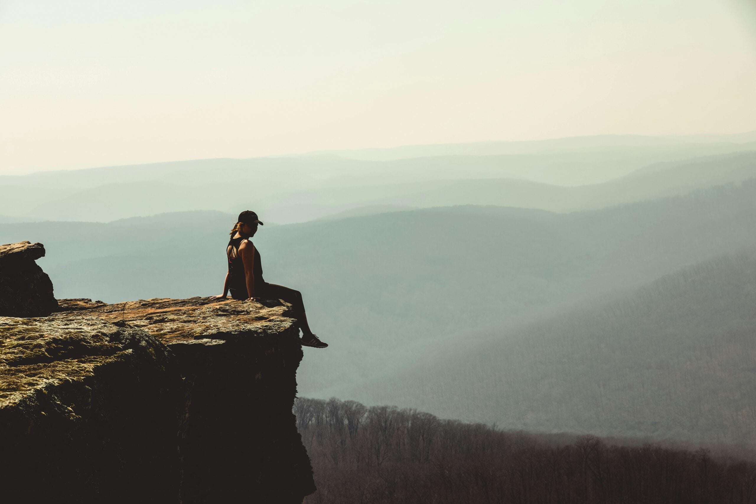 A person wearing a cap sits on the edge of a rocky cliff, overlooking a vast, hazy landscape of rolling hills and valleys under a clear sky.