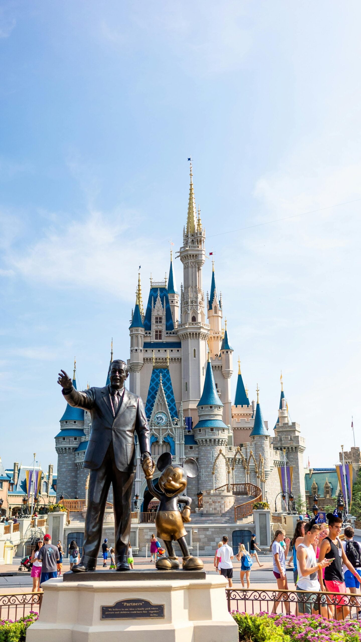 A bronze statue of a man holding hands with Mickey Mouse stands in front of a large, ornate castle at a busy theme park, with visitors walking around under a clear blue sky.