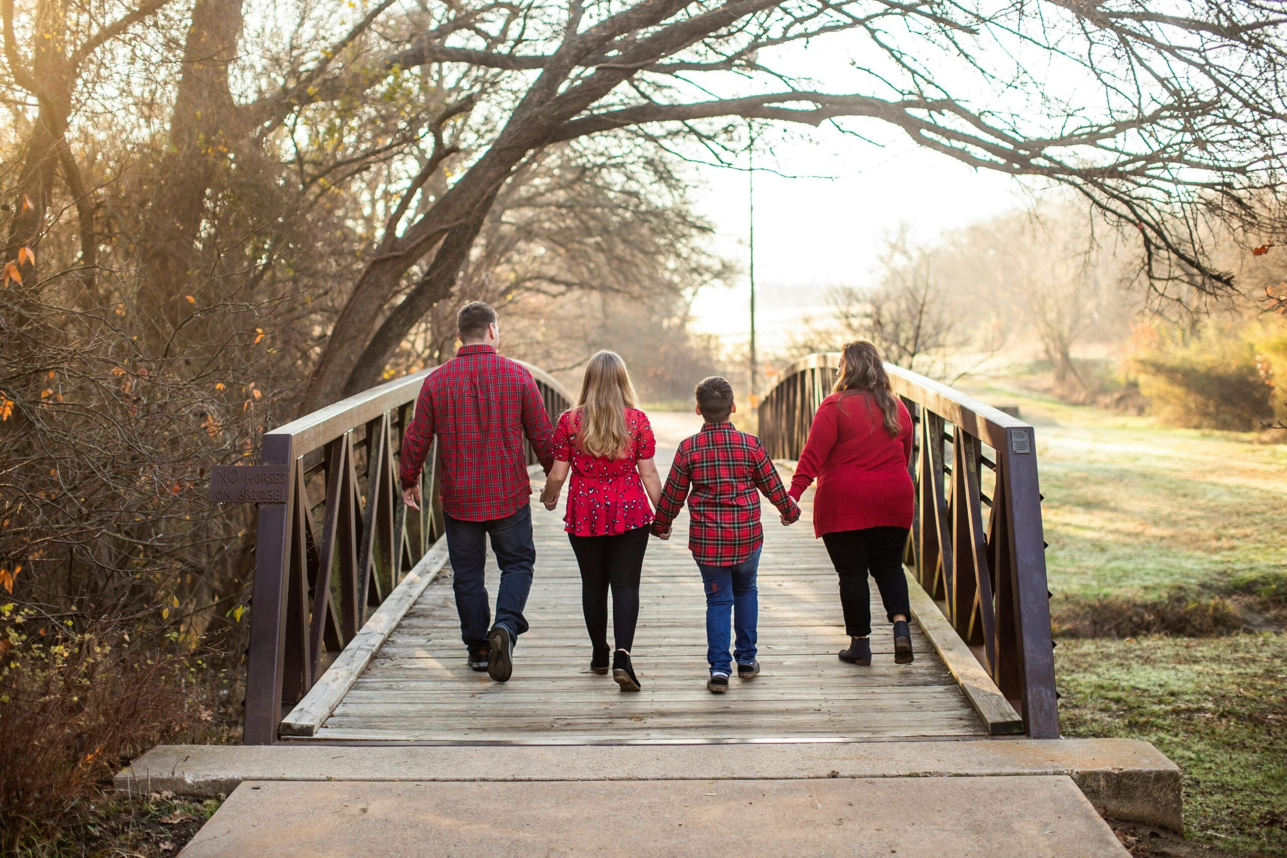 Four people, two adults and two children, walk hand-in-hand away from the camera on a wooden bridge surrounded by trees, all wearing red tops in a natural, outdoor setting.