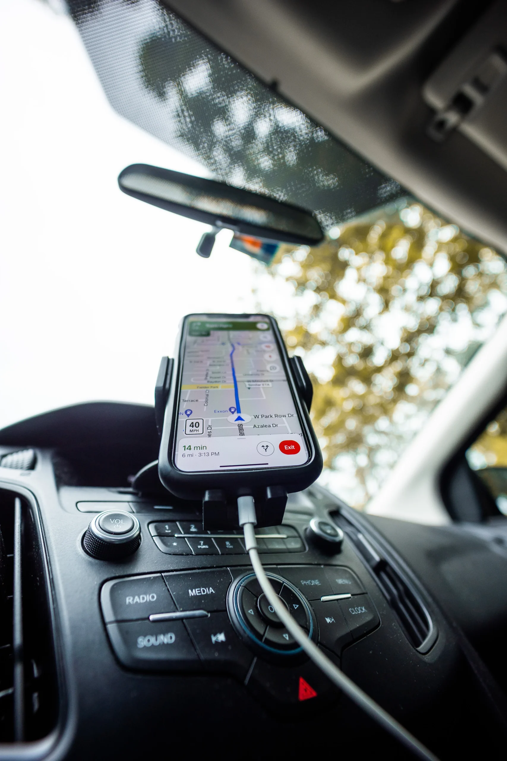 A smartphone displaying a GPS navigation map is mounted on a car dashboard, connected by a charging cable, with trees visible through the windshield in the background.