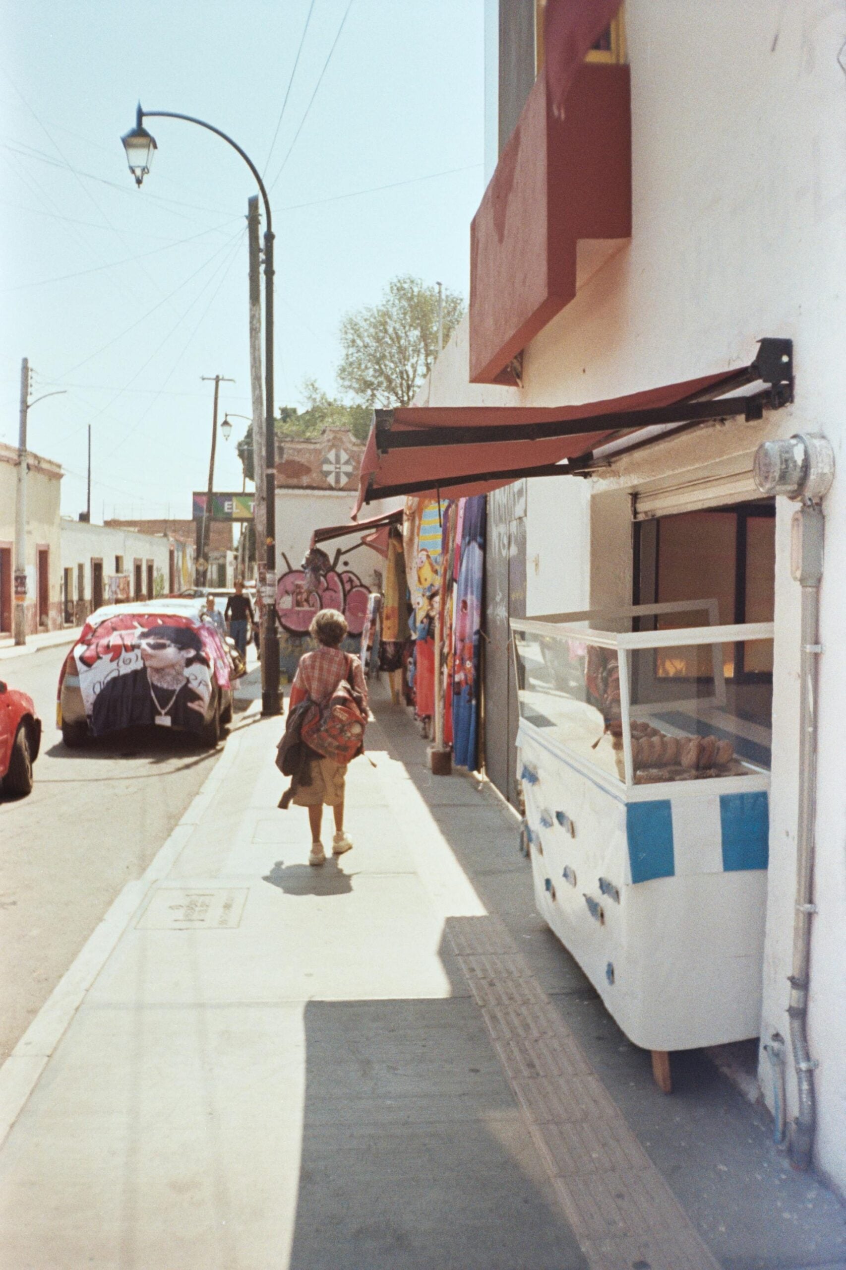 A woman with a backpack and a child walk down a sunny sidewalk lined with shops and a glass display case; colorful fabric covers a parked car nearby.