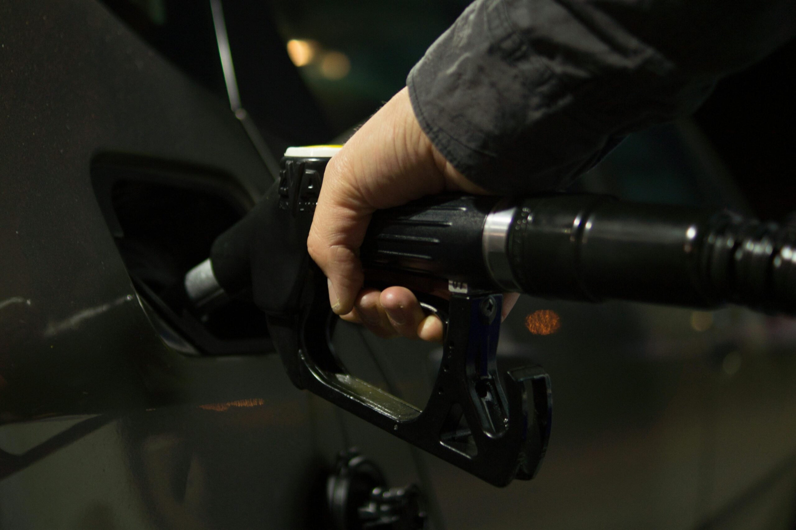 A person’s hand holds a black fuel pump nozzle, refueling a car at a gas station. The nozzle is inserted into the car’s fuel tank, and the background is dimly lit.