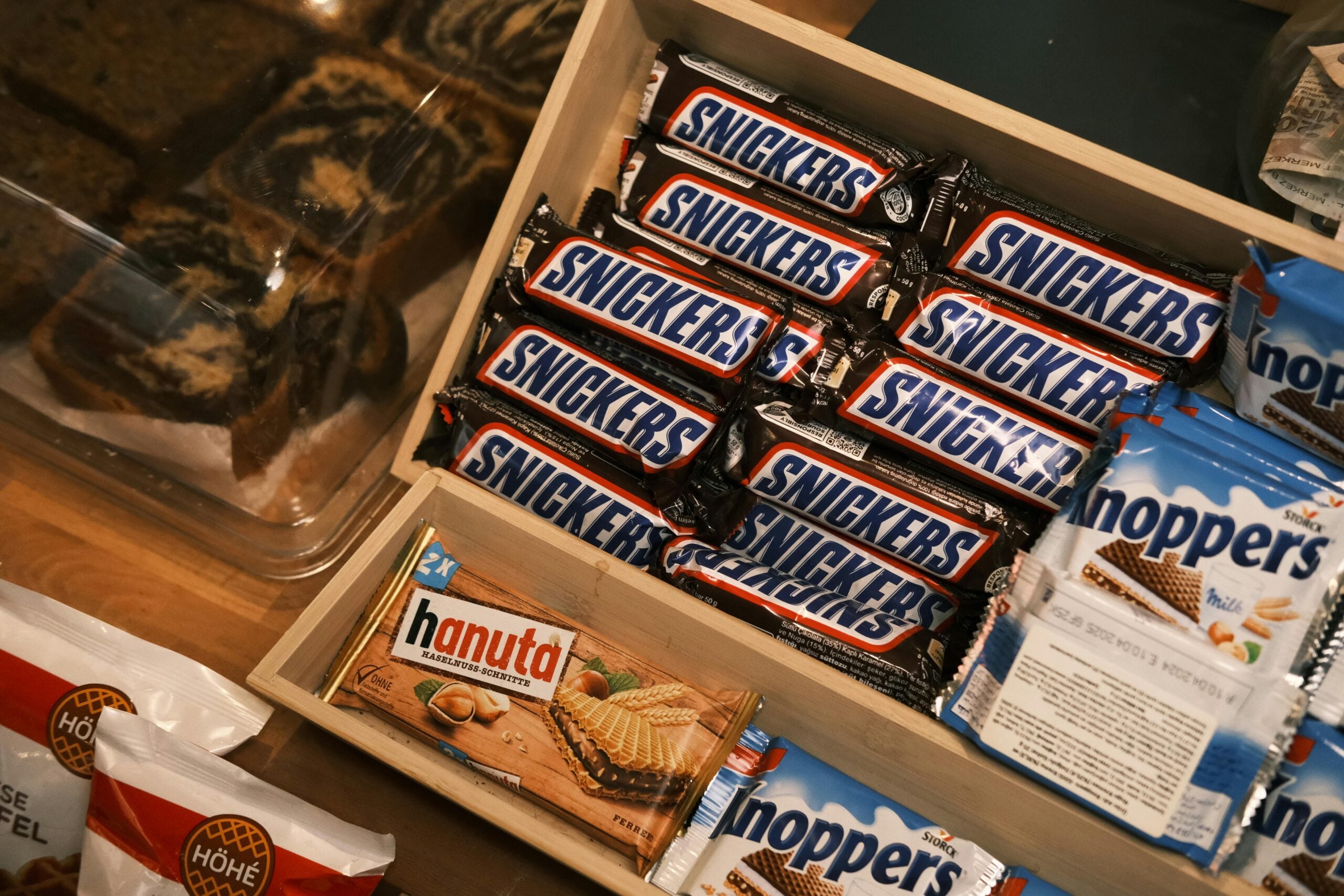 An open wooden box filled with multiple Snickers bars, a Hanuta wafer snack, and several Knoppers wafer snacks, all on a wooden surface with other food items partially visible nearby.