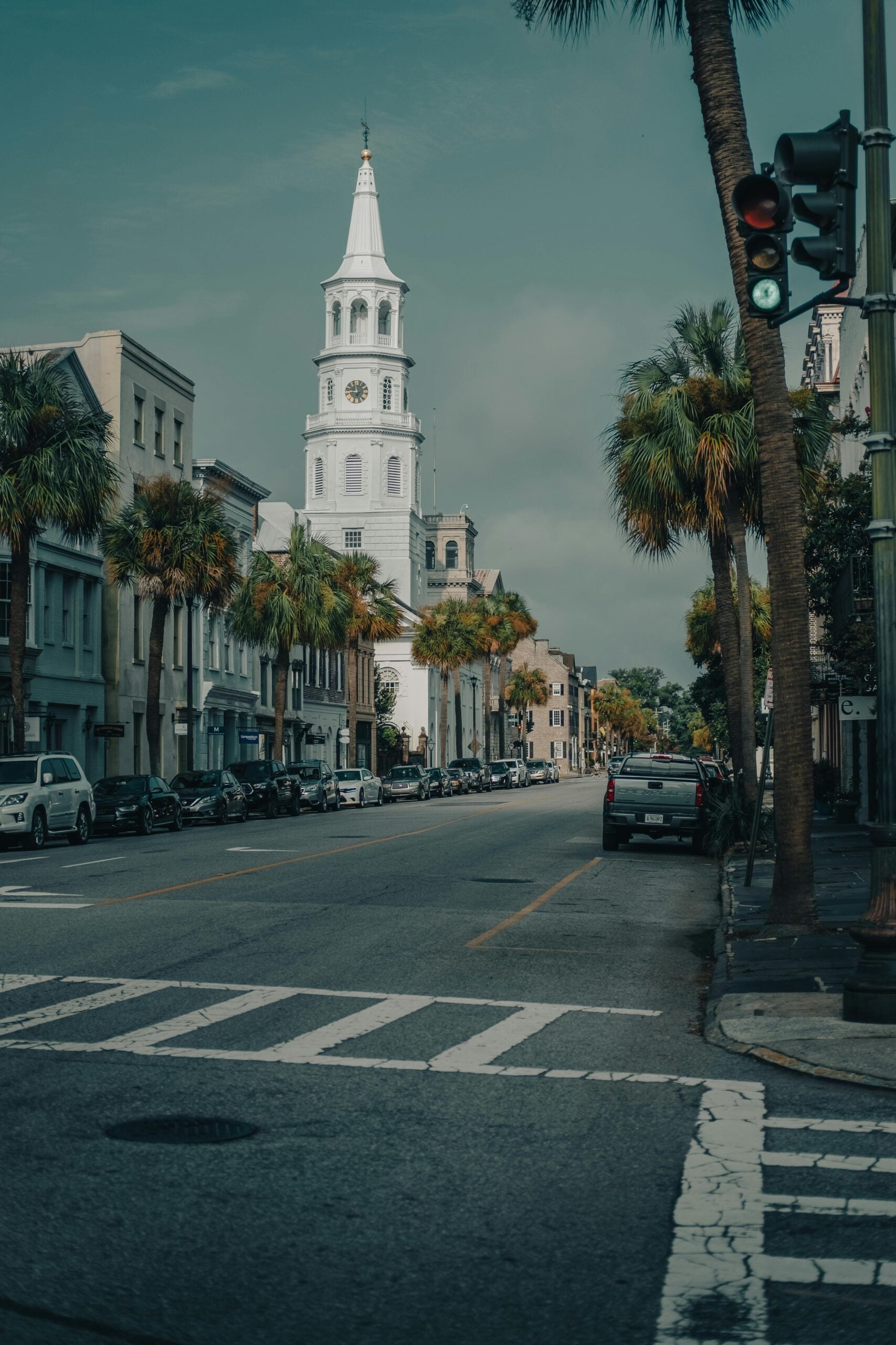 Walkable towns in America: A city street lined with palm trees and parked cars, featuring a tall white church steeple with a clock, under a partly cloudy sky and a green traffic light in the foreground.