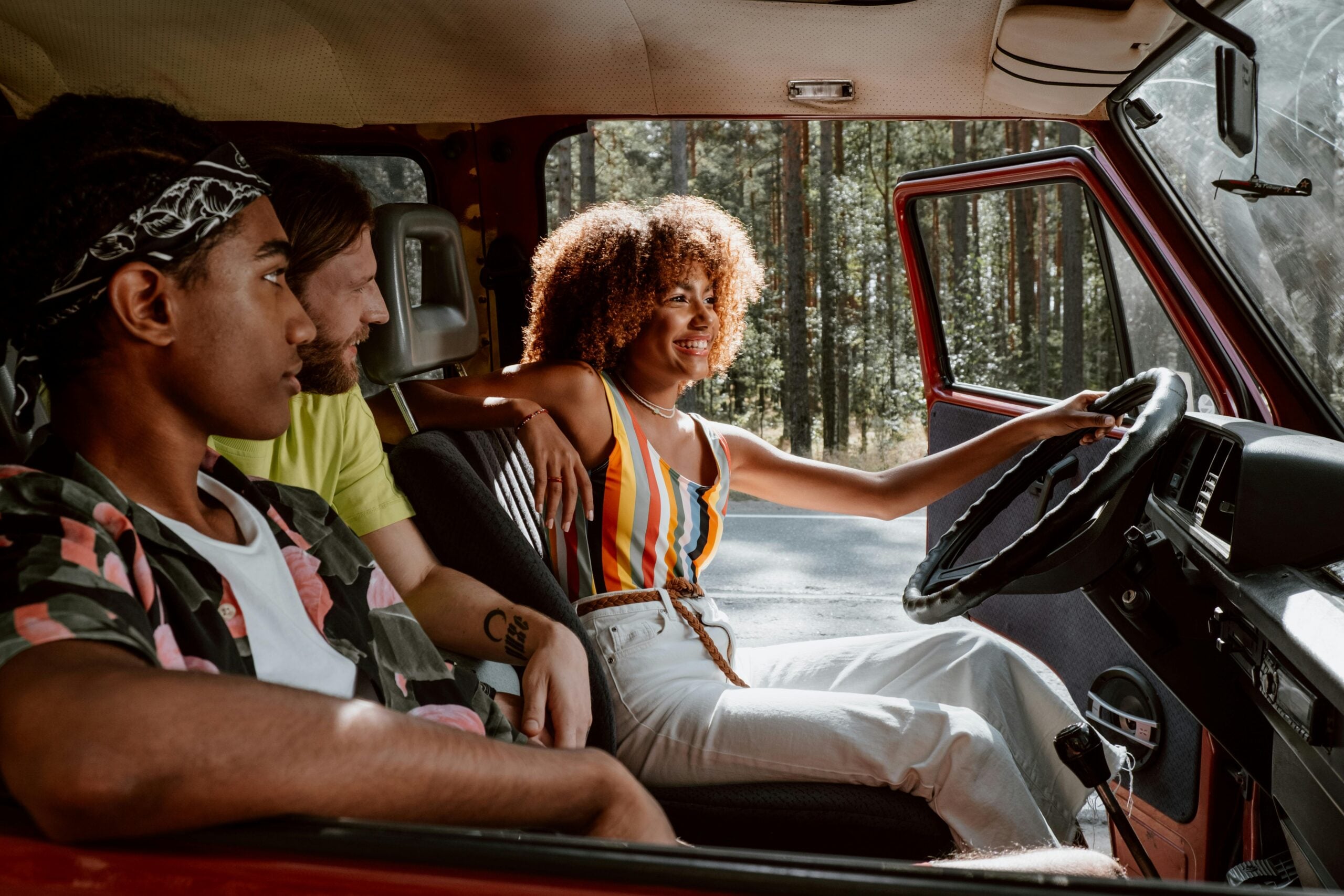 Three friends are sitting in a car; a woman in colorful clothes smiles while driving, and two men sit beside her, looking relaxed. Sunlight comes through the windows and trees are visible outside.