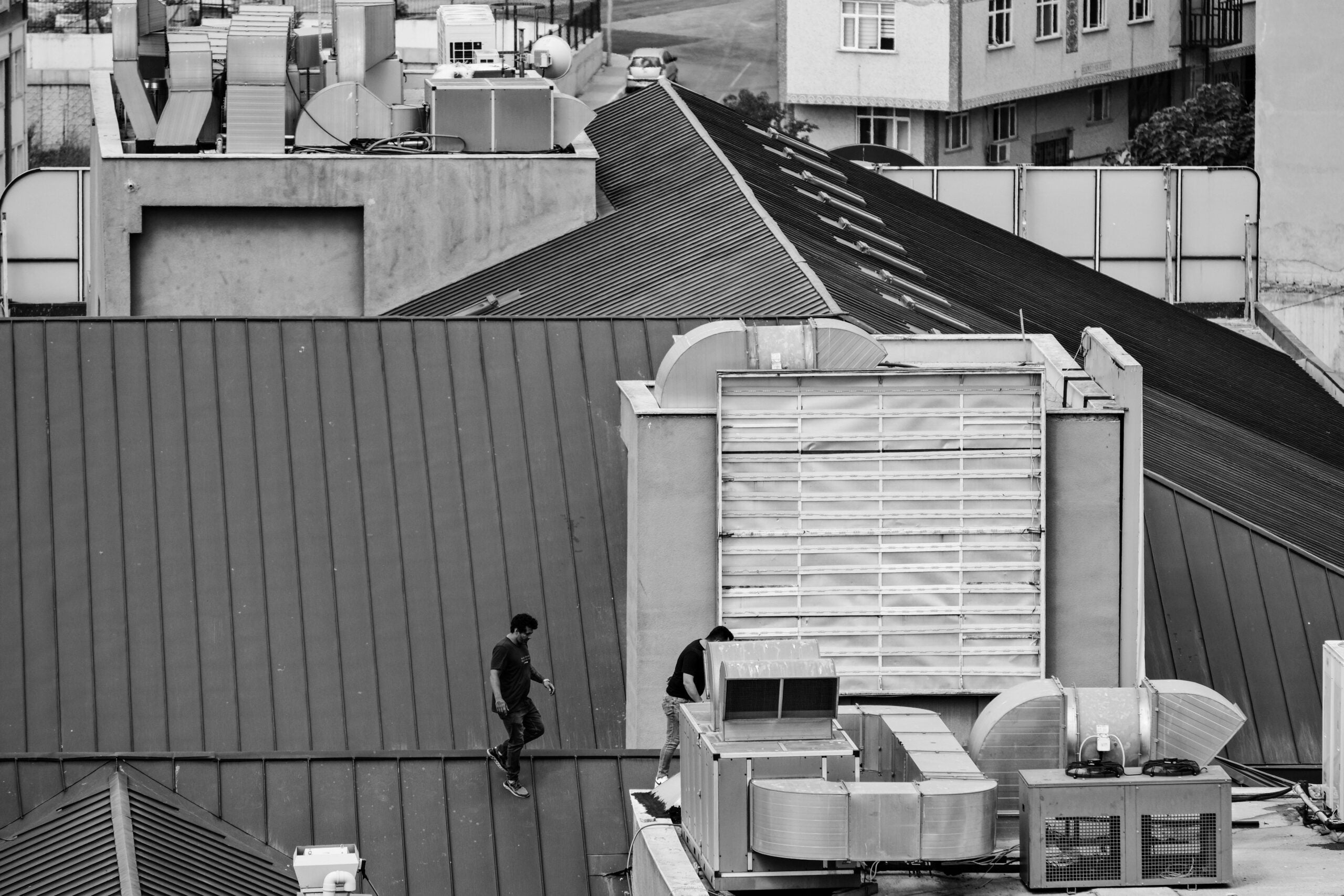 Two people stand on an industrial rooftop surrounded by large HVAC units and ventilation systems; nearby buildings are visible in the background. The image is black and white.