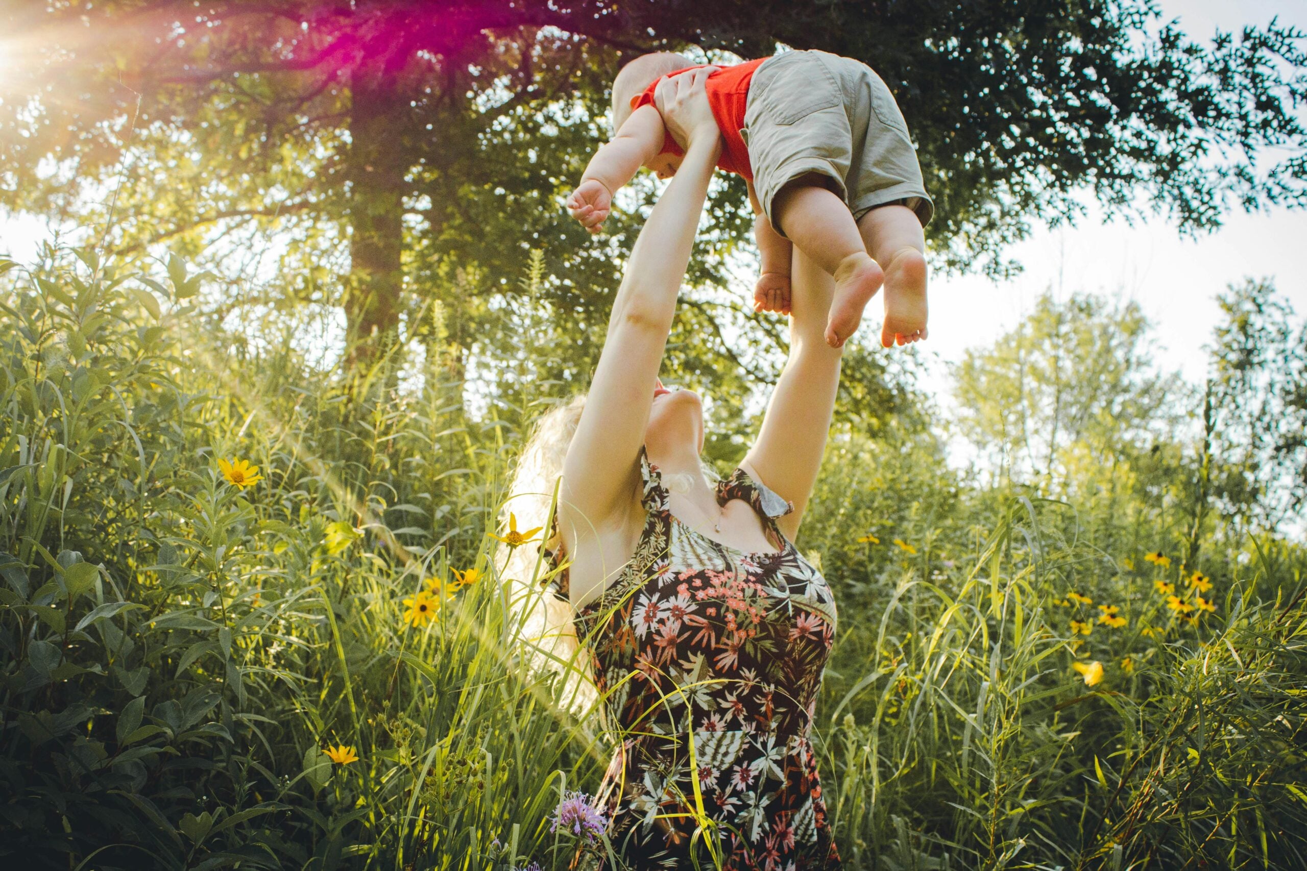 A woman in a floral dress stands in tall grass and wildflowers, lifting a baby above her head on a sunny day, with sunlight streaming through the trees in the background.