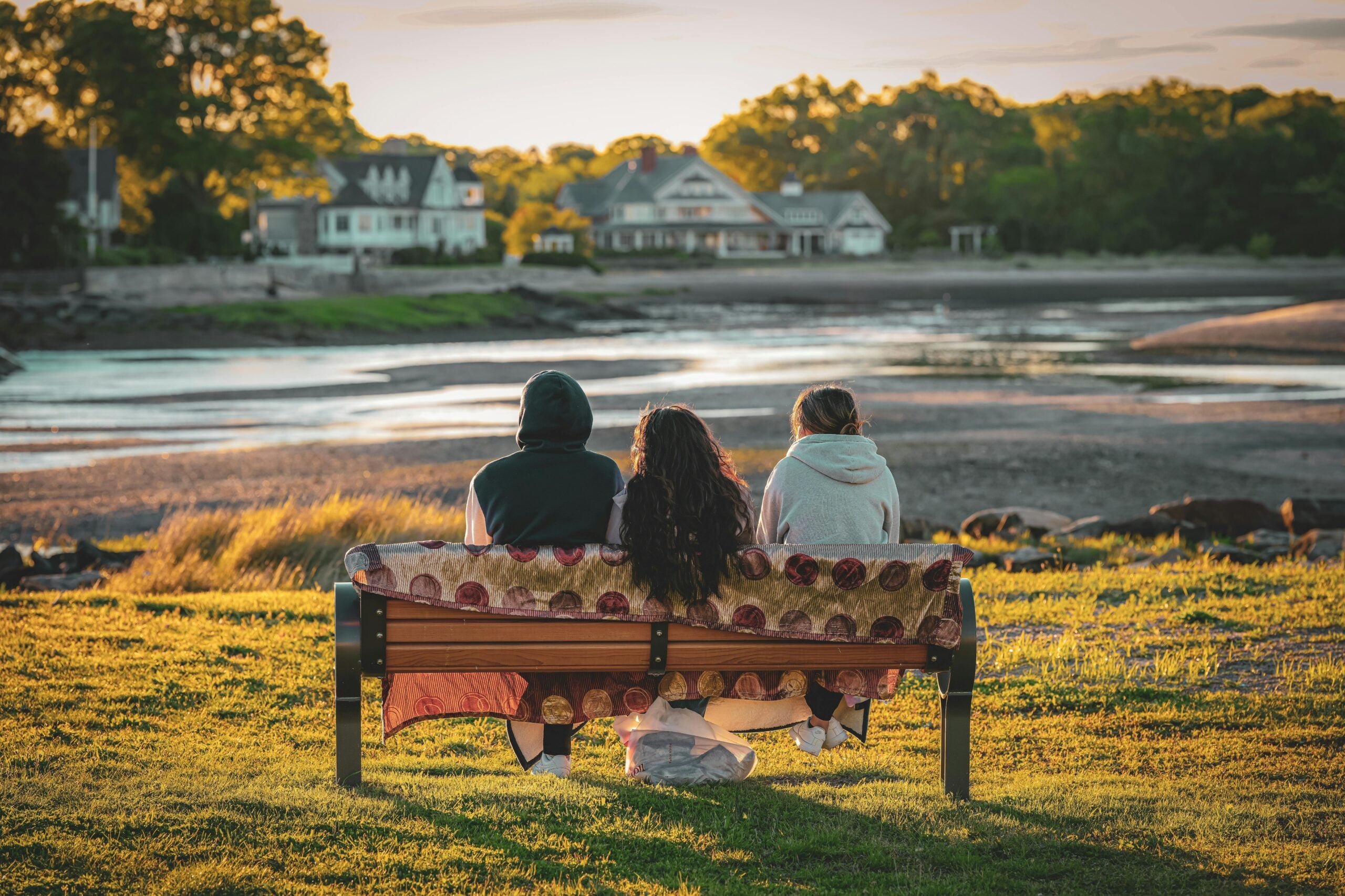 Three people sit on a bench, facing a scenic riverside view with houses and trees in the background, as warm sunlight illuminates the grass and water.