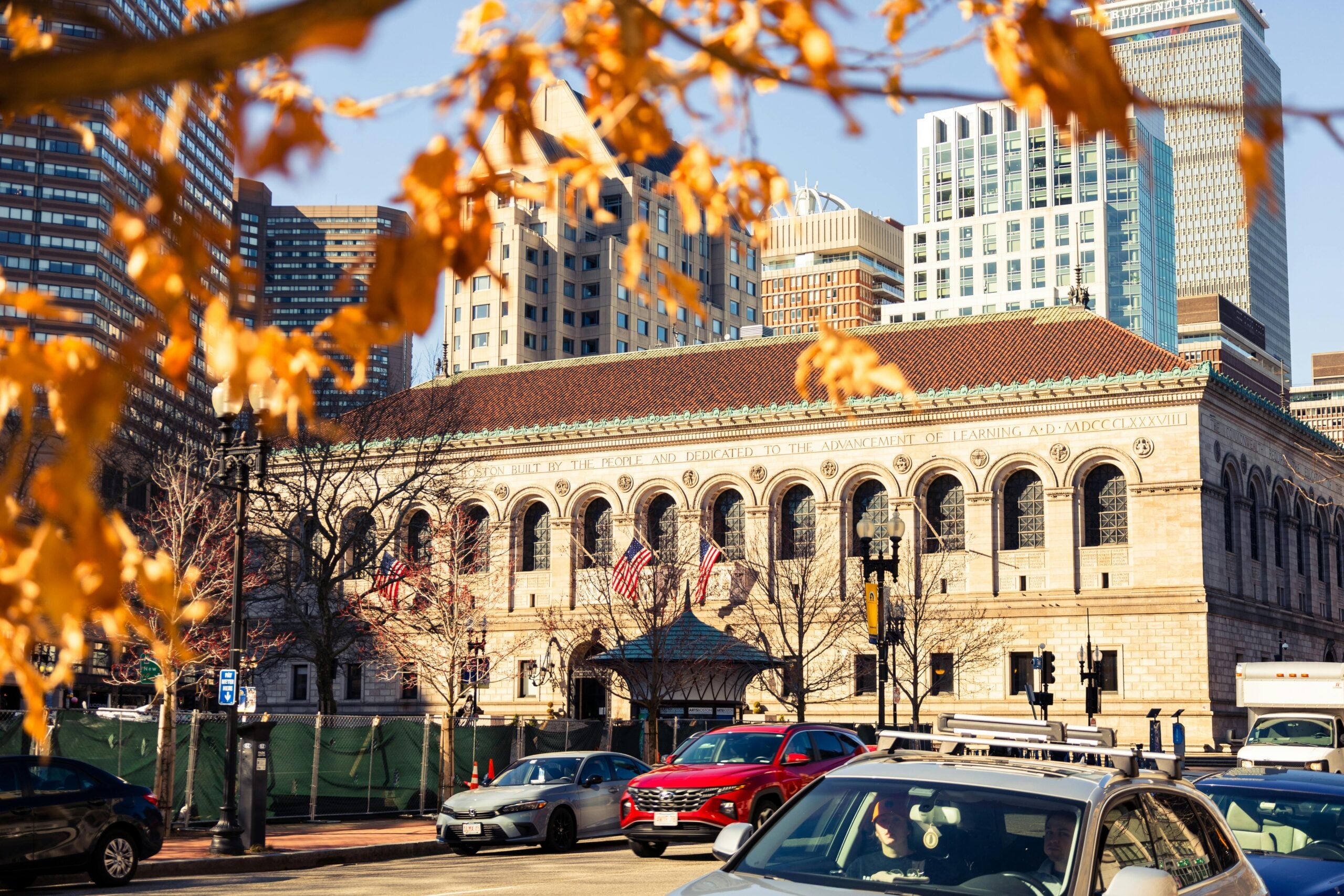 The Boston Public Library building with American flags at the entrance, surrounded by city skyscrapers, autumn leaves in the foreground, and cars parked on the street in bright sunlight.