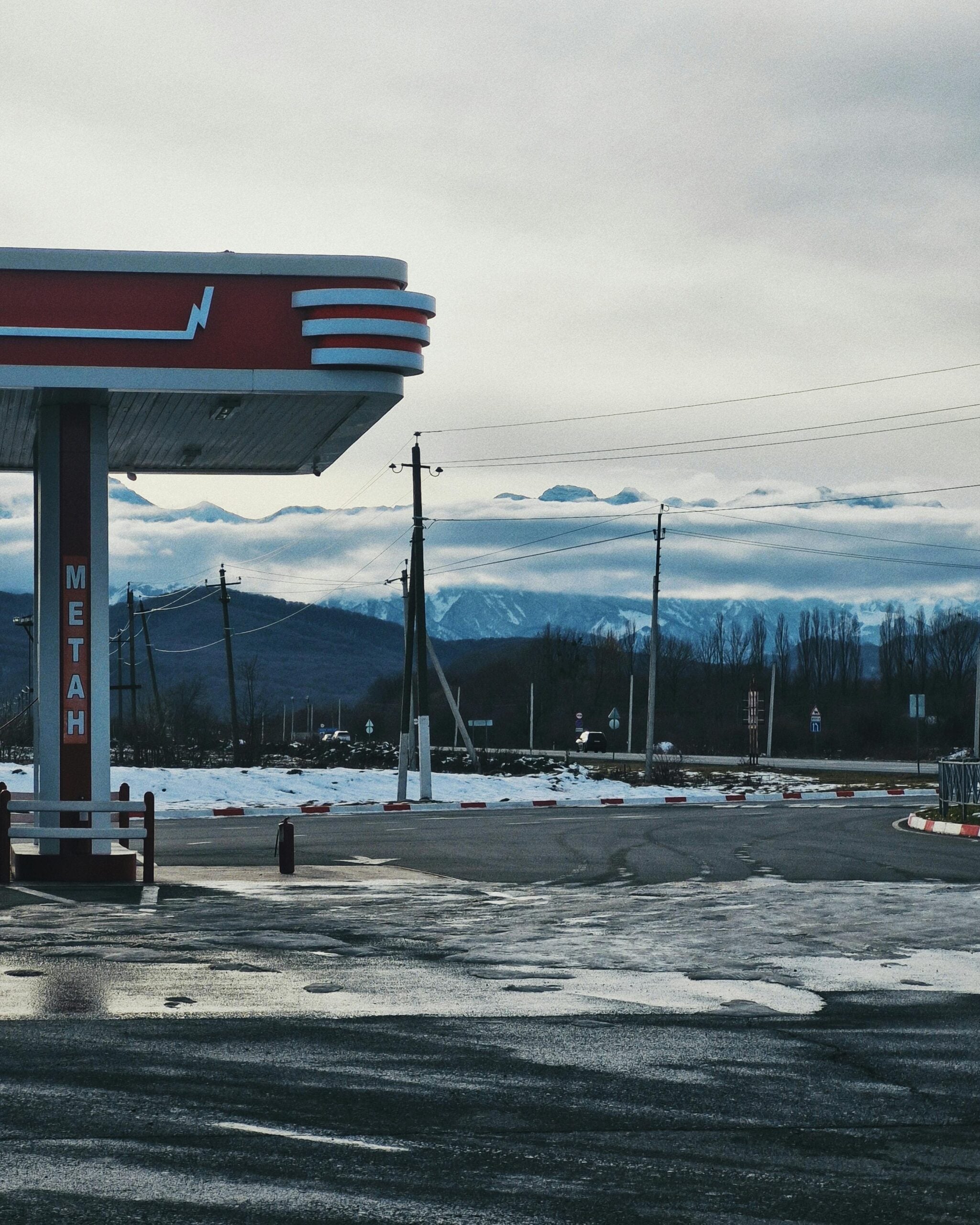A gas station with a red and white canopy stands on a wet, icy pavement. Snow is scattered on the ground, and mountains are visible in the distance under a cloudy sky. Utility poles and wires line the roadside.