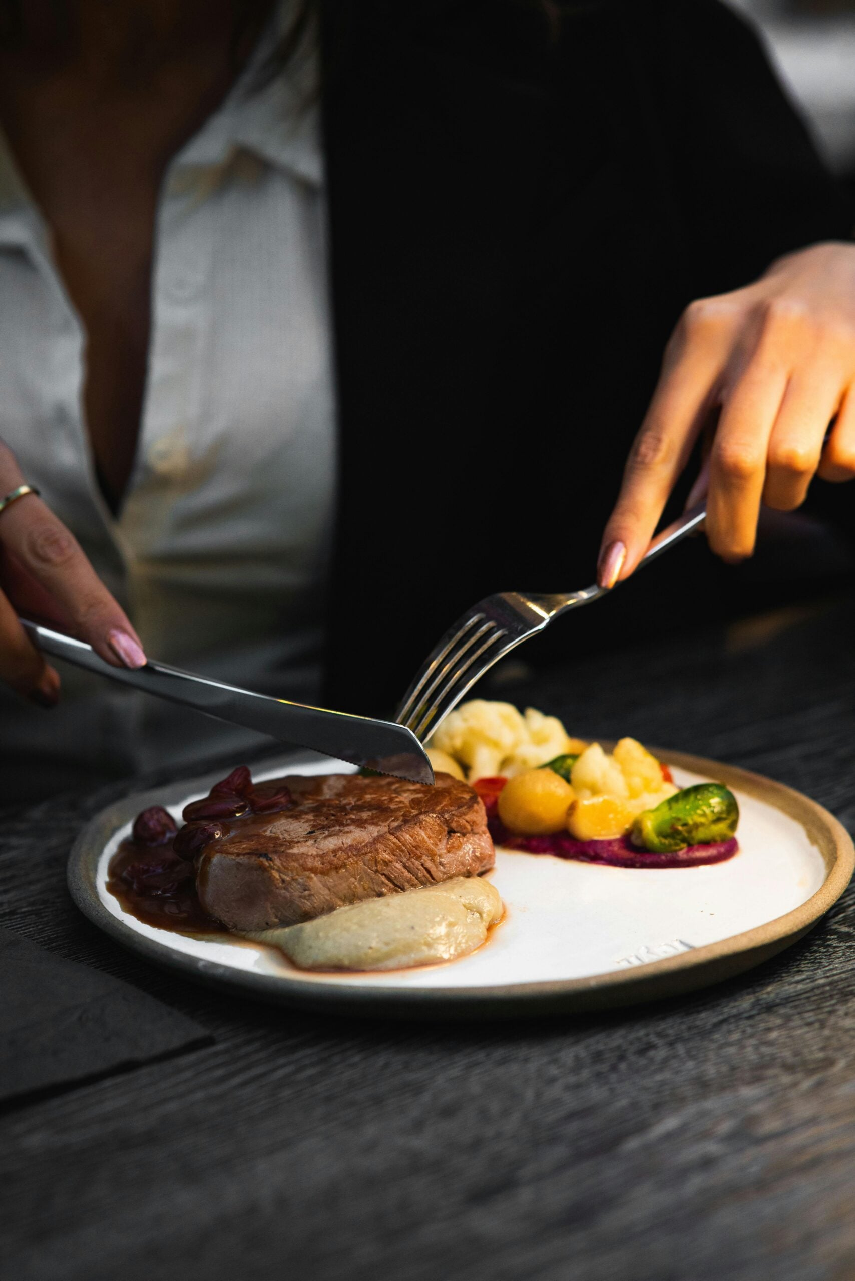 A person cuts into a steak on a plate with a fork and knife. The plate also has mashed potatoes, vegetables, and sauce, and the person is wearing a white shirt and black jacket.