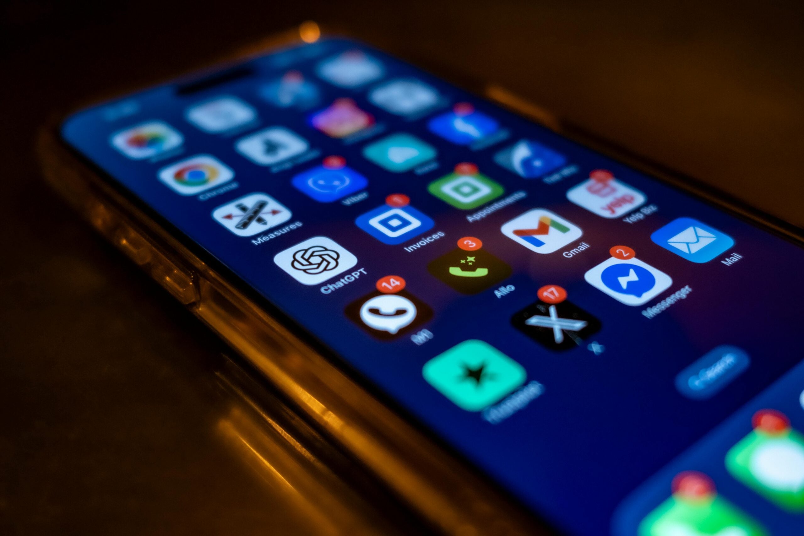 A close-up of a smartphone screen displaying various app icons, some with notification badges. The phone is placed on a dark surface, and the image is slightly angled, focusing on the lower half of the screen.