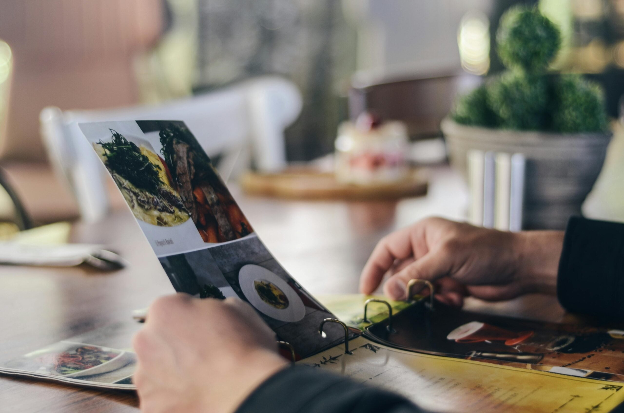 A person browses a restaurant menu at a wooden table, with blurred plates, a plant, and soft lighting in the background.